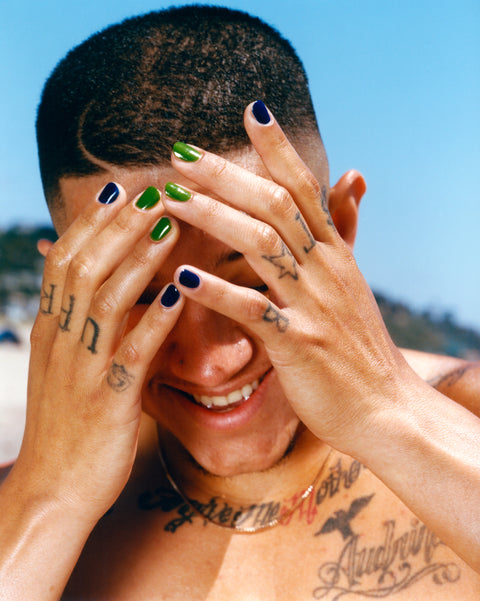 Man with green and navy painted nails holding his hands in front of his face.